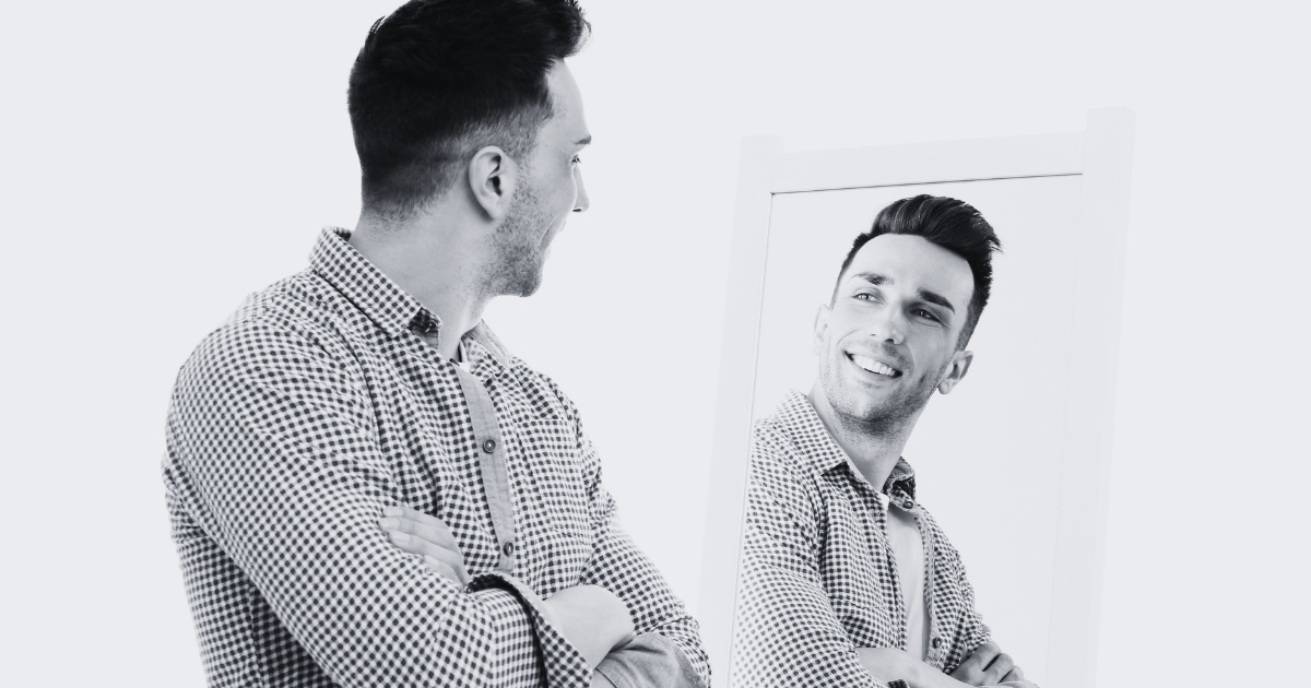 A man wearing a checkered shirt stands with his arms crossed, looking into a mirror and smiling at his own reflection.