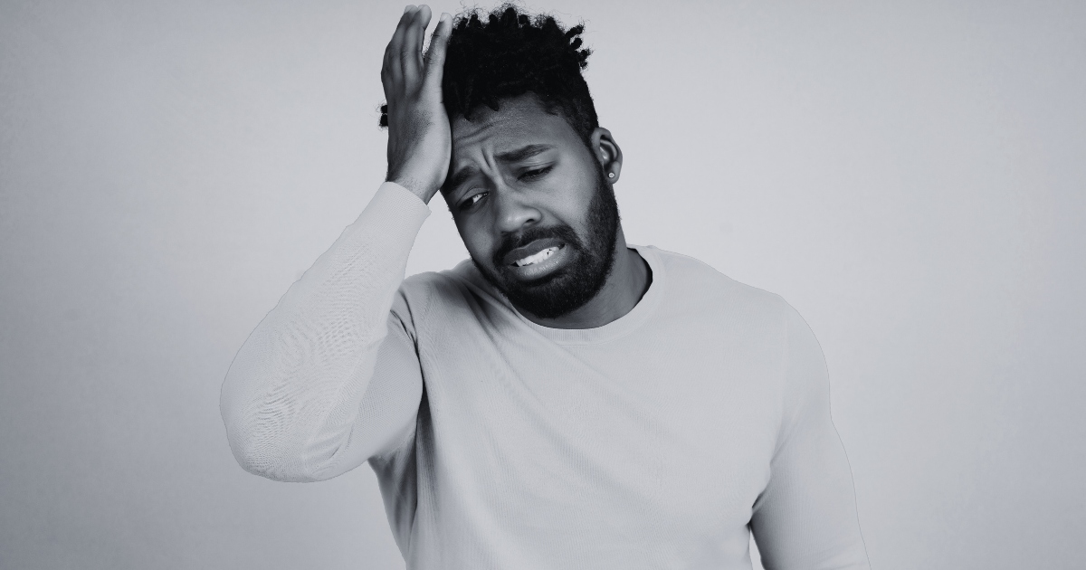 A man with a beard wearing a light-colored long-sleeve shirt looks distressed, holding his hand to his head with a pained expression on his face.