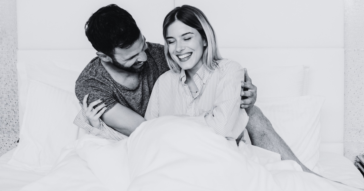 A black-and-white photo of a couple sitting together in bed, smiling and laughing. The man has his arm around the woman, and they appear happy and relaxed.